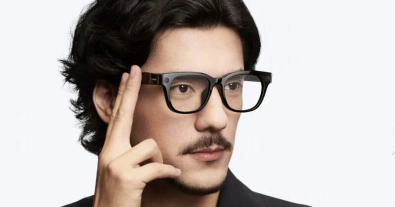 Un hombre con cabello negro y bigote usa gafas inteligentes y toca el costado del marco con los dedos, como si usara un control táctil. El fondo es simple y de color claro.