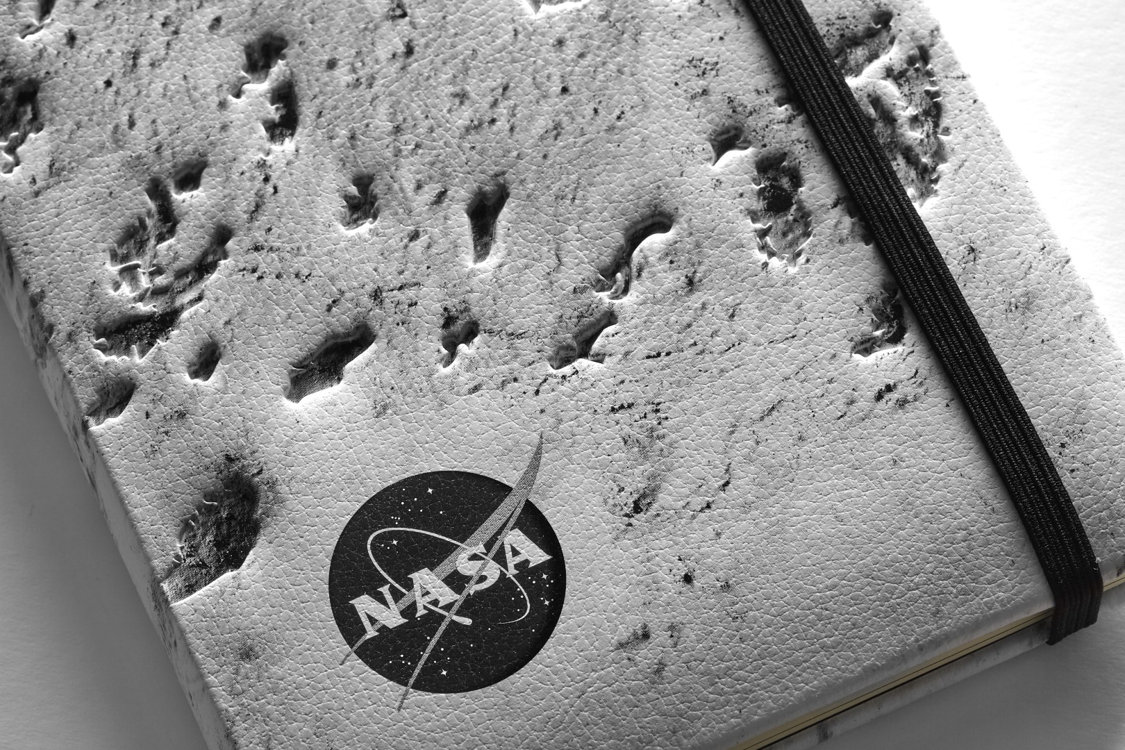Cierre un cuaderno de edición limitada inspirada en la NASA por Moleskine en plata. La cubierta es un cierre de la superficie de la luna. El cuaderno tiene un cierre elástico negro. Una versión en blanco y negro del logotipo de la NASA está en la esquina inferior izquierda.