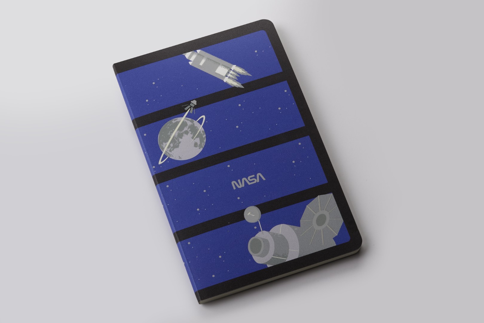 Soft Coor Journal de la colección de cuadernos de edición limitada inspirada en la NASA por Moleskine. Hay cuatro paneles azul oscuro en una columna, cada uno con su propia imagen de escala de grises. De arriba a abajo: cohete, tierra con satélite en órbita, texto de la NASA, satélite grande.