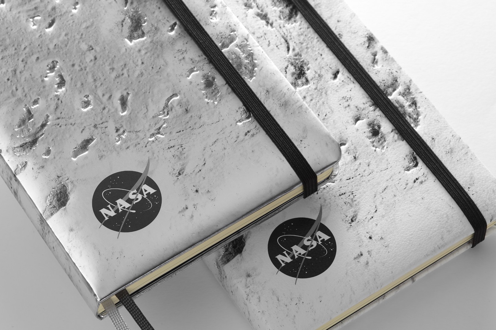 Dos cuadernos de edición limitada inspirados en la NASA de Moleskine en plata oscura y plata. Cada cubierta es un cierre de la superficie de la luna, tiene un cierre elástico negro y una versión en blanco y negro del logotipo de la NASA está en la esquina inferior izquierda.