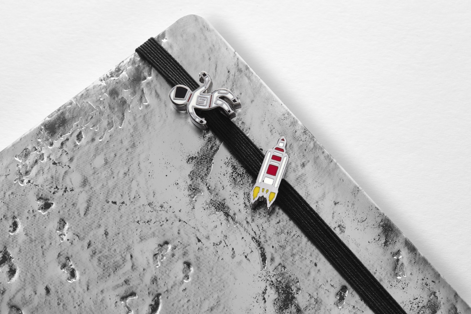 El primer piso de dos pines pequeños unidos al cierre elástico negro del cuaderno de edición limitada inspirada en la NASA por Moleskine (superficie de la luna plateada). El pivote superior es una persona con un vestido de astronauta blanco con acabado rojo. El pivote inferior es un pequeño cohete simplista gris, blanco y rojo que explota.