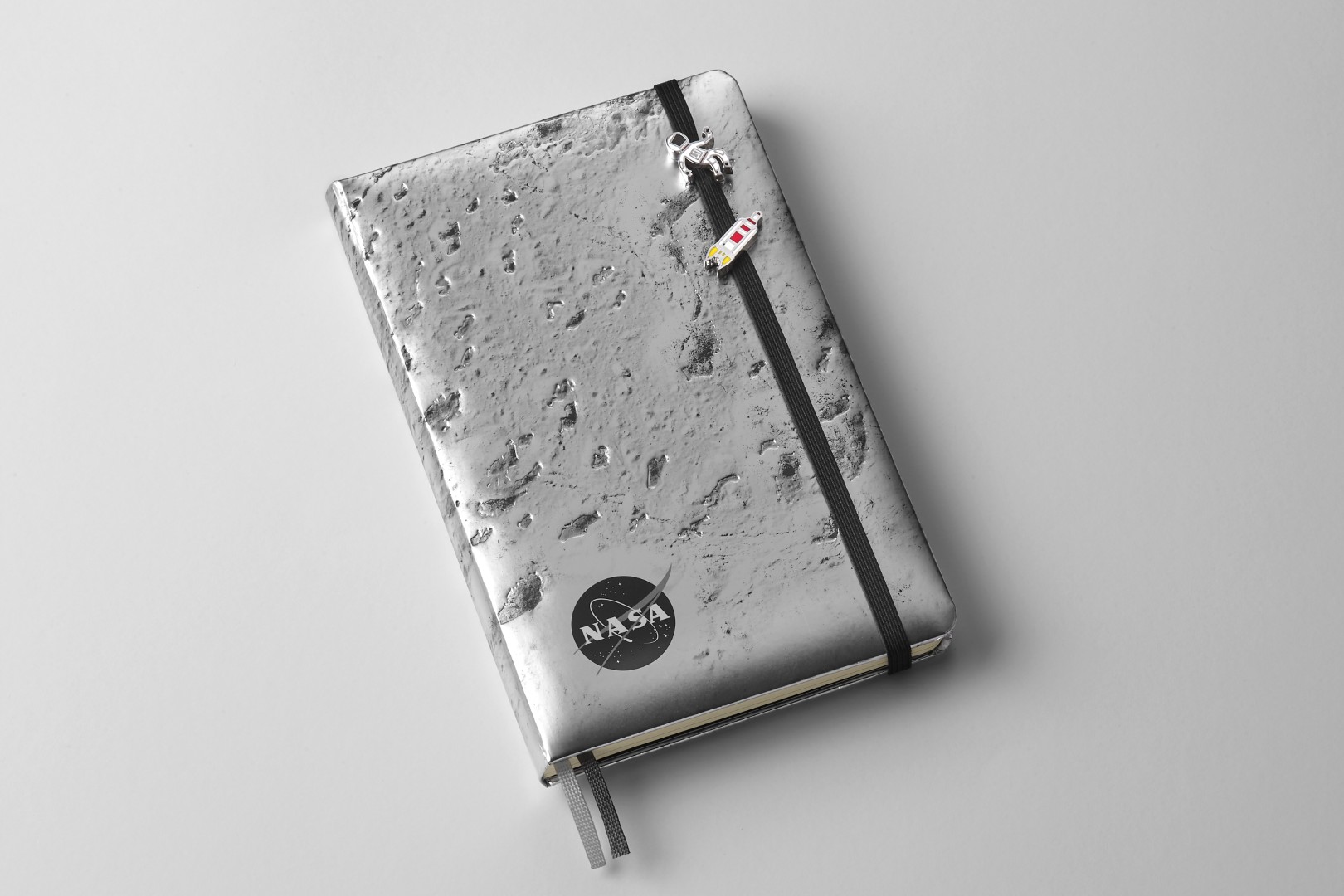 Dos pines pequeños unidos al cierre elástico negro del cuaderno de edición limitada inspirados en la NASA por Moleskine (superficie de la luna plateada). El pivote superior es una persona con un vestido de astronauta blanco con acabado rojo. El pivote inferior es un pequeño cohete simplista gris, blanco y rojo que explota.