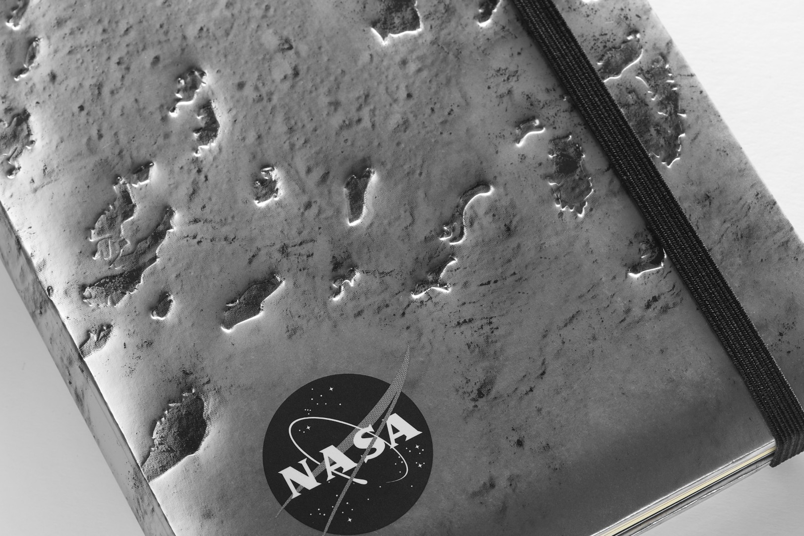 Cierre un cuaderno de edición limitada inspirada en la NASA por Moleskine en plata oscura. La cubierta es un cierre de la superficie de la luna. El cuaderno tiene un cierre elástico negro. Una versión en blanco y negro del logotipo de la NASA está en la esquina inferior izquierda.