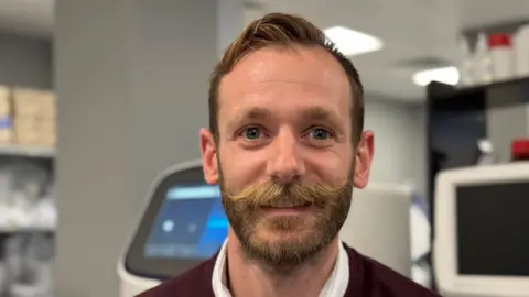 BBC Un hombre con barba y grandes sonrisas de bigote a la cámara. Está parado en un laboratorio.