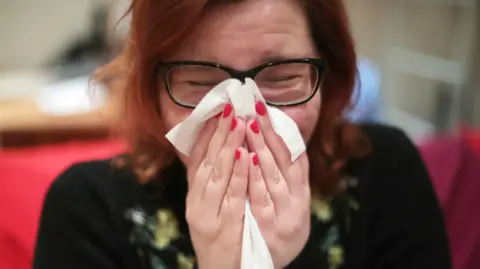 Un medio cercano a una mujer con cabello rojo y uñas pintadas en rojo que se soplan la nariz con una tela. Sus ojos están cerrados detrás de las gafas con marco oscuro. Un sofá rojo y un escritorio de madera se han borrado detrás de ella.