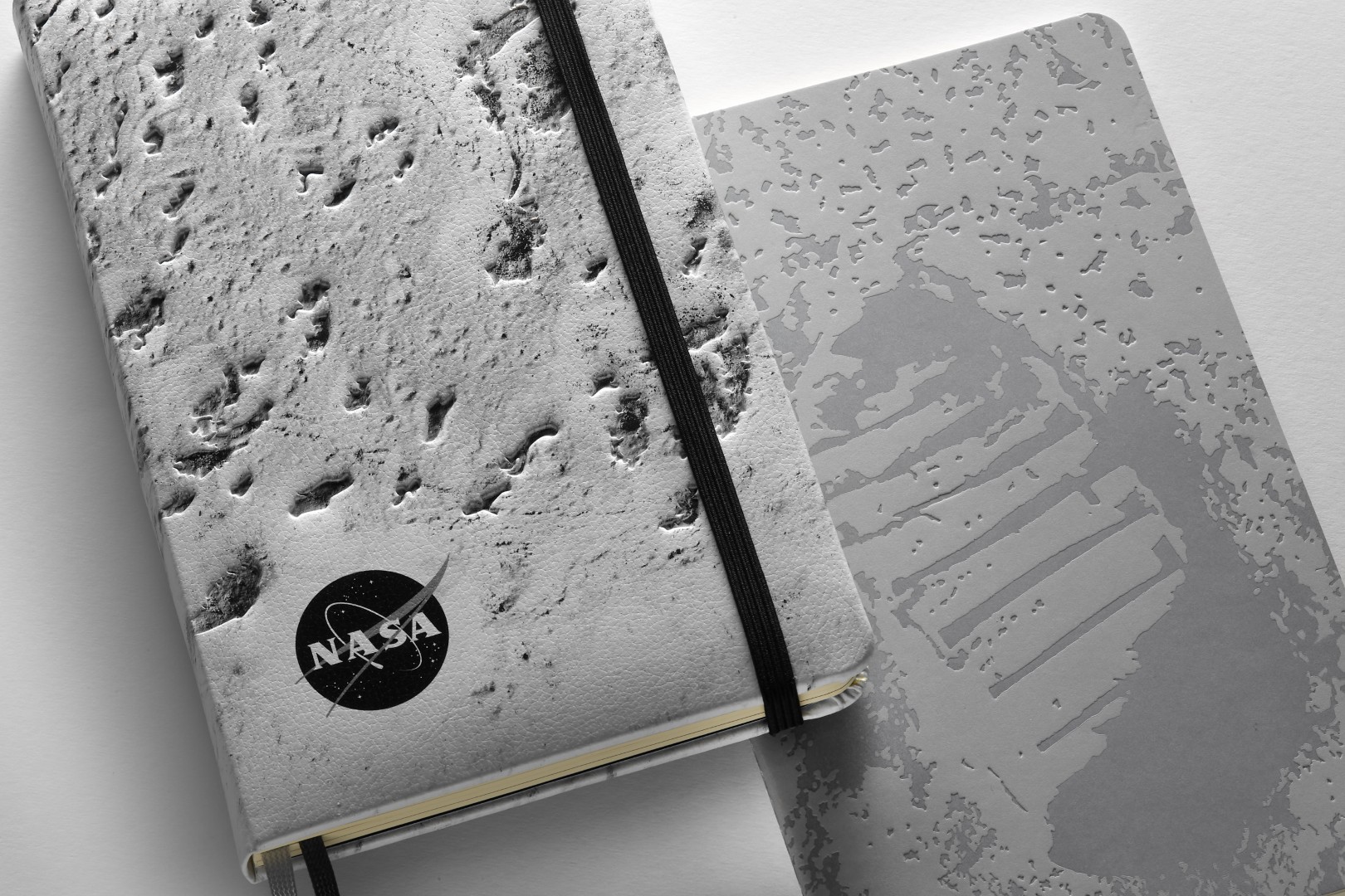 Notebook de edición limitada inspirada en la NASA de Moleskine. Izquierda: cuaderno con cubierta rígida con el primer piso de la superficie de la luna en la cubierta delantera, logotipo de la NASA en blanco y negro en la cubierta en la parte inferior izquierda. A la derecha: cobertura suave con la huella de Neil Armstrong en la luna en la portada.