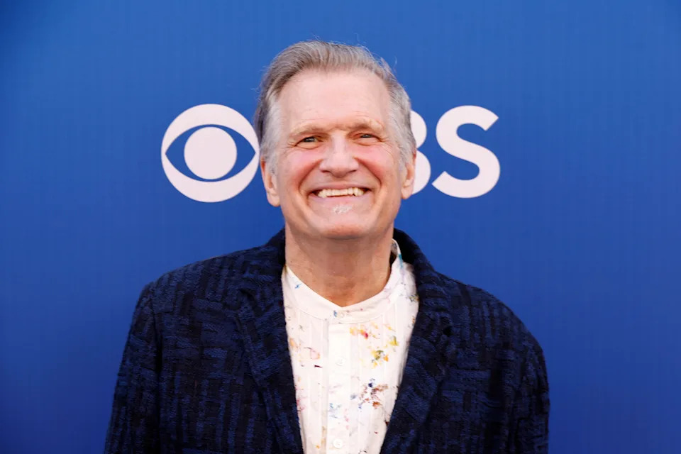 Una persona con una chaqueta estampada sonríe frente a un fondo con el logo de CBS, probablemente en un evento para los medios.