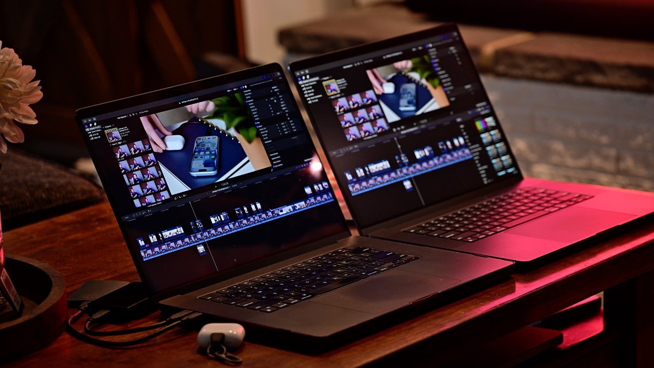Dos portátiles MacBook Pro de 16 pulgadas sobre una mesa de madera que muestran un software de edición de vídeo. Un estuche pequeño y una vista parcial de una flor blanca cercana. Iluminación cálida al fondo.