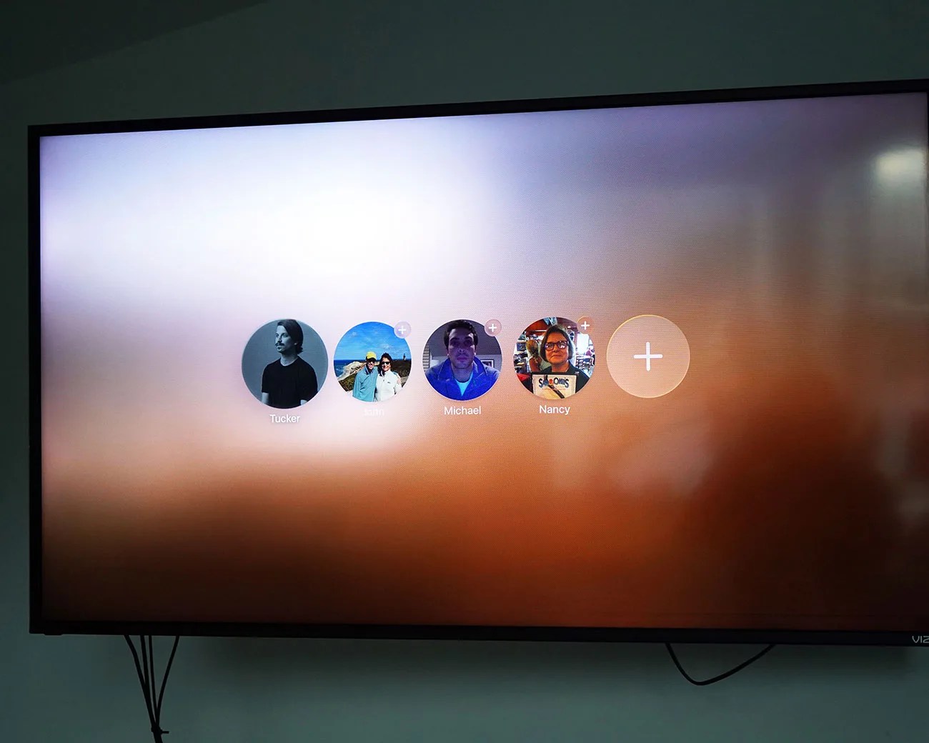 Pantalla de televisión que muestra un menú de selección de perfil de usuario con cinco íconos circulares etiquetados como Tucker, Sam, Michael, Nancy y un signo más para agregar un perfil.