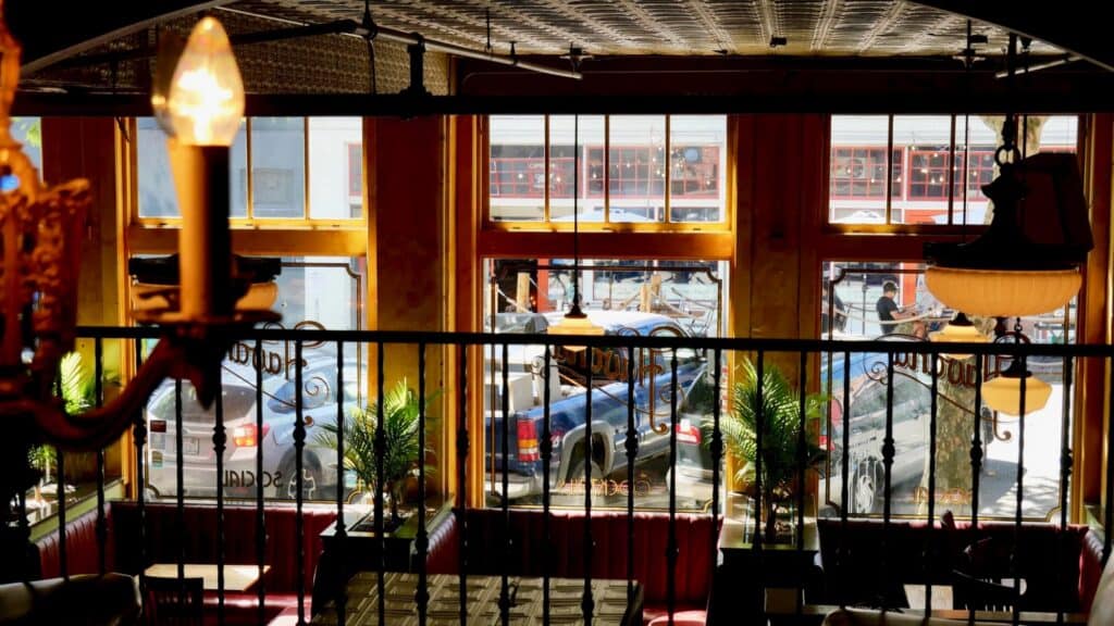 Vista desde el interior de un restaurante con grandes ventanales, cabinas rojas, plantas decorativas y autos estacionados visibles afuera en un día soleado.